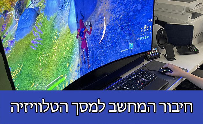 אדם המשתמש במחשב המחובר למסך טלוויזיה מעוקל. המסך מציג משחק וידאו, ויש מכשירים אלקטרוניים שונים על השולחן. הטקסט בתחתית הוא בעברית.