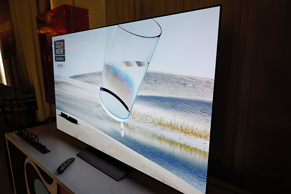טלוויזיה בעלת מסך שטוח מציגה תמונה של כוס מים מקרוב על נוף מדברי. שלט רחוק מונח על המעמד מתחת לטלוויזיה. על תווית הטלוויזיה נכתב "2024 NEW QD-OLED 55/65/77".