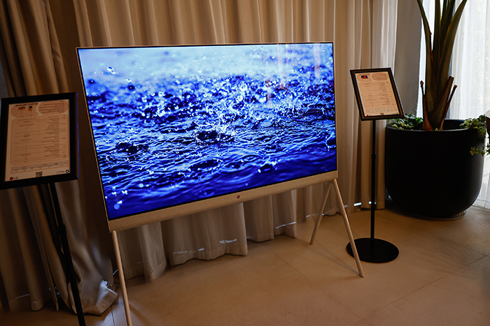טלוויזיה בעלת מסך שטוח המציגה תמונה של טיפות מים ניתזות, כששני מעמדים מחזיקים ניירות מכל צד וברקע עציץ.