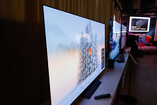 טלוויזיה מודרנית בעלת מסך שטוח המציגה תמונה של עץ מכוסה שלג בחדר אפלולי, כשברקע רואים טלוויזיה נוספת ושלט רחוק.