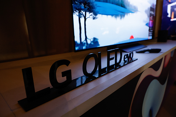 שלט שחור "LG OLED evo" מוצג על דלפק מול מסך גדול המציג סצנת טבע עם עצים ומים.